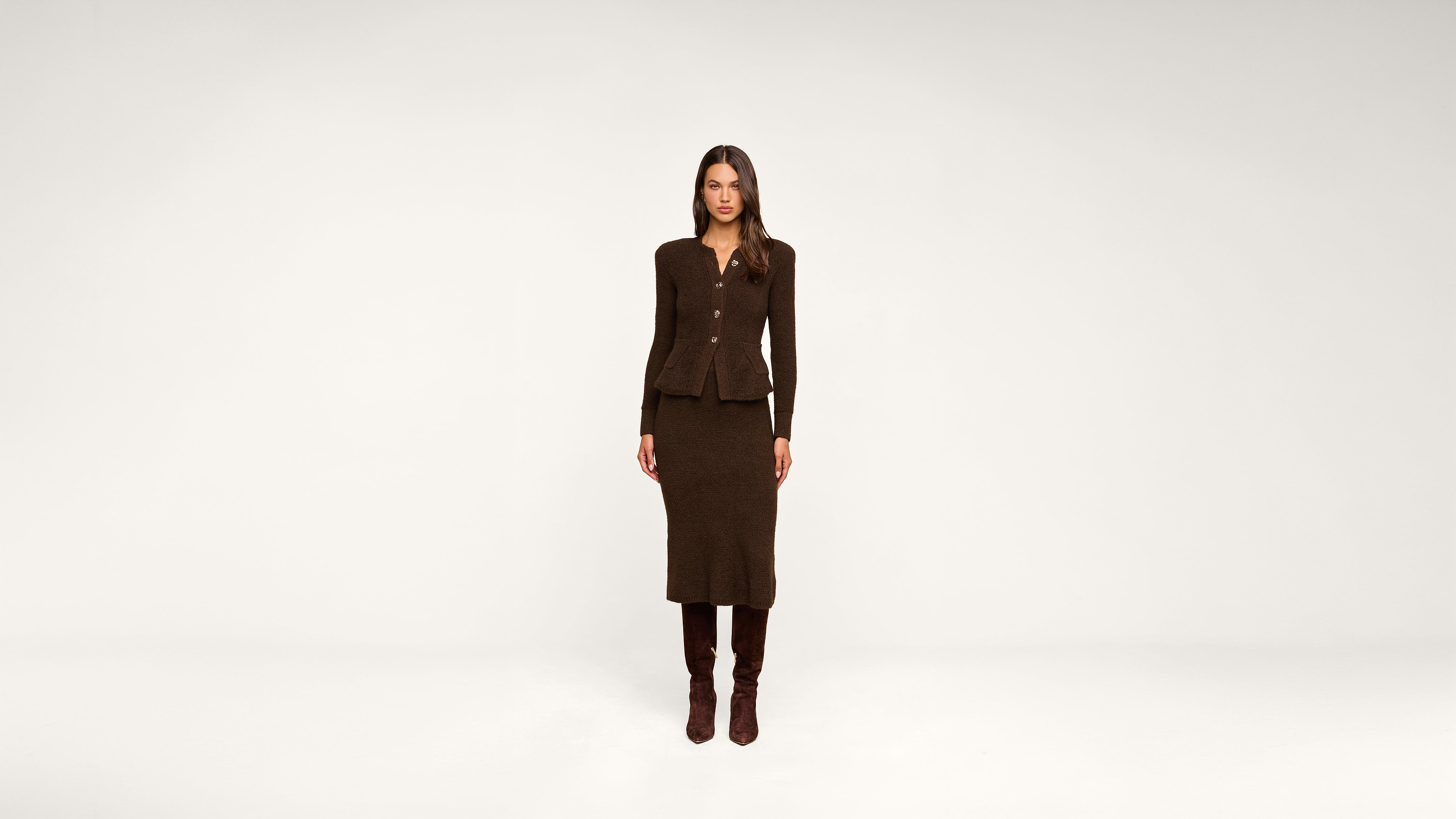 A woman stands facing forward in a brown, buttoned jacket and matching skirt with knee-high brown boots, against a plain white background.