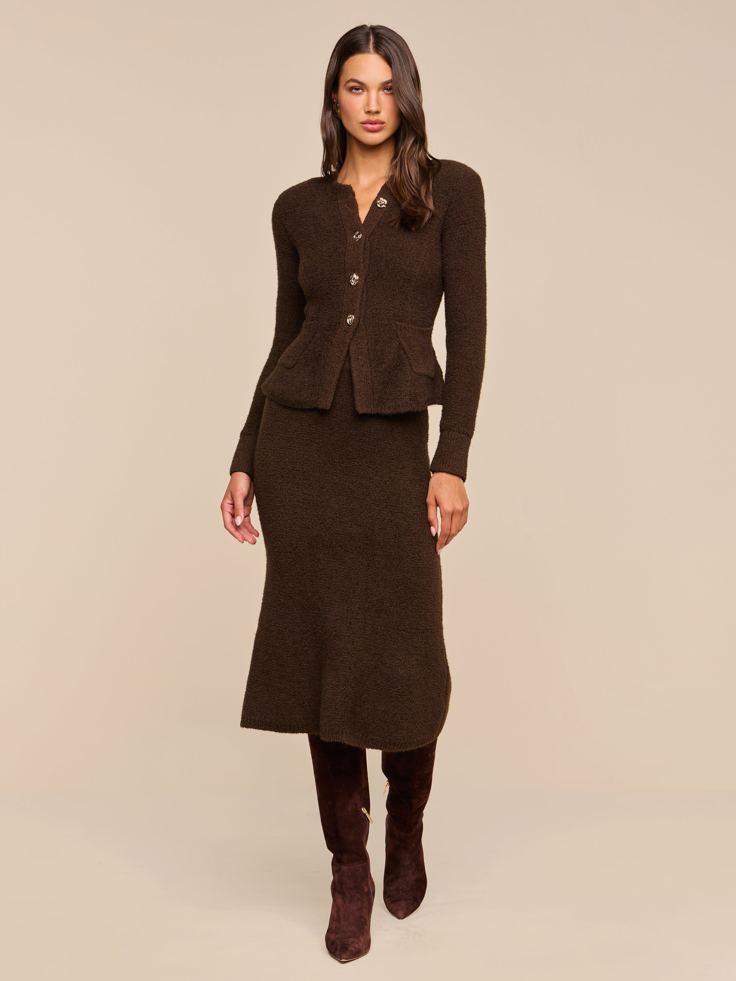 Woman wearing the Melissa Knit Cardigan—a fitted, dark brown blazer crafted from textured knit yarn with button details—paired with a matching midi skirt and knee-high brown boots, standing against a plain beige background for an elegant take on women’s knitwear.