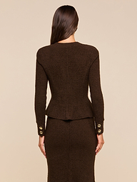 A woman with long brown hair, seen from behind, is wearing the Melissa Knit Cardigan—a dark brown textured knit reminiscent of sophisticated women’s knitwear—paired with a matching skirt. The cardigan features gold buttons on the sleeve cuffs. The background is beige.