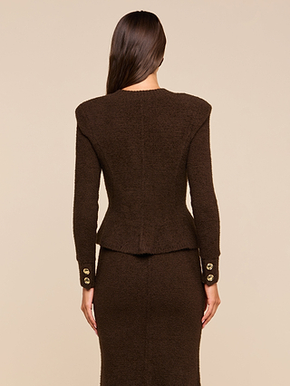 A woman with long brown hair, seen from behind, is wearing the Melissa Knit Cardigan—a dark brown textured knit reminiscent of sophisticated women’s knitwear—paired with a matching skirt. The cardigan features gold buttons on the sleeve cuffs. The background is beige.