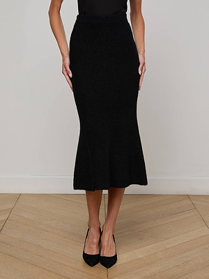 A woman in the L'AGENCE Salem Knit Skirt—black, fitted with a slight flare and elastic waistband—pairs it with a black sleeveless top and pointed-toe heels, standing on a light wood floor against a plain wall.
