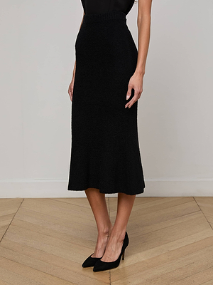 A woman models the L'AGENCE Salem Knit Skirt, featuring a black design and elastic waistband, paired with a black sleeveless top and high heels. She stands on a light wood floor against a plain wall, with her face not visible.