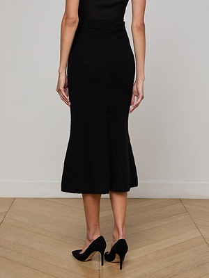 A woman stands indoors on a wooden floor, shown from behind, wearing the L'AGENCE Salem Knit Skirt—a black, high-waisted skirt with a slight flare and elastic waistband—paired with a black sleeveless top and black heels.