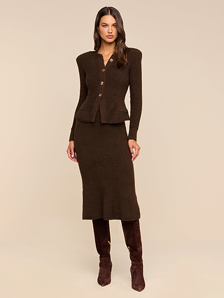 A woman with long hair and a neutral expression stands against a beige background, wearing a dark brown long-sleeve buttoned top, knee-high brown boots, and the Salem Knit Skirt—a matching soft knit midi skirt.
