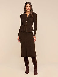 A woman stands against a beige background wearing the Salem Knit Skirt, a fitted, dark brown midi skirt with a soft, textured knit finish. She pairs it with a matching buttoned cardigan and dark brown knee-high boots.