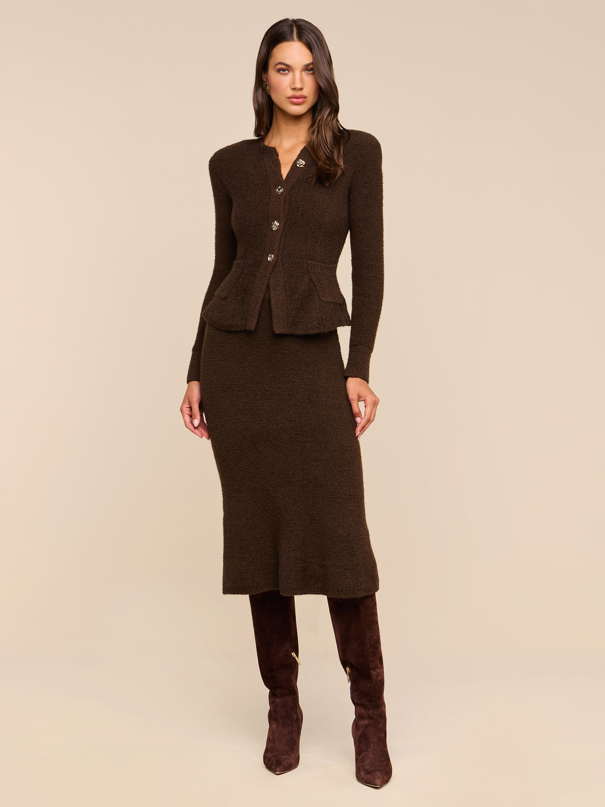 A woman stands against a beige background wearing the Salem Knit Skirt, a fitted, dark brown midi skirt with a soft, textured knit finish. She pairs it with a matching buttoned cardigan and dark brown knee-high boots.