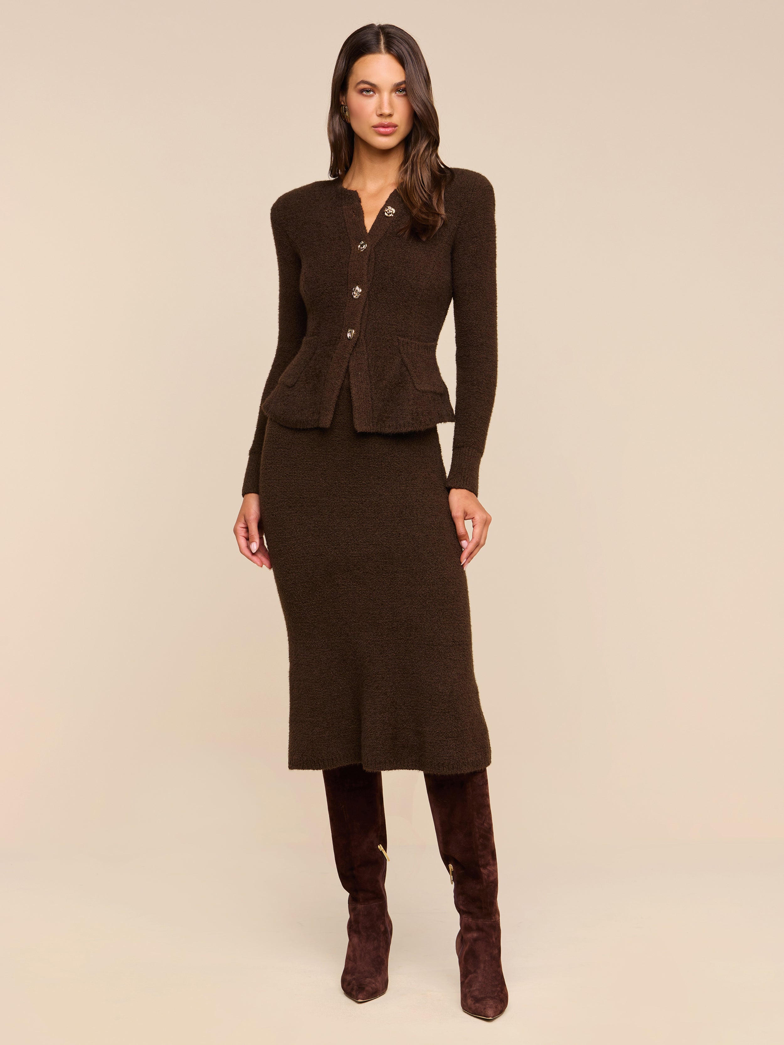 A woman stands against a beige background wearing the Salem Knit Skirt, a fitted, dark brown midi skirt with a soft, textured knit finish. She pairs it with a matching buttoned cardigan and dark brown knee-high boots.