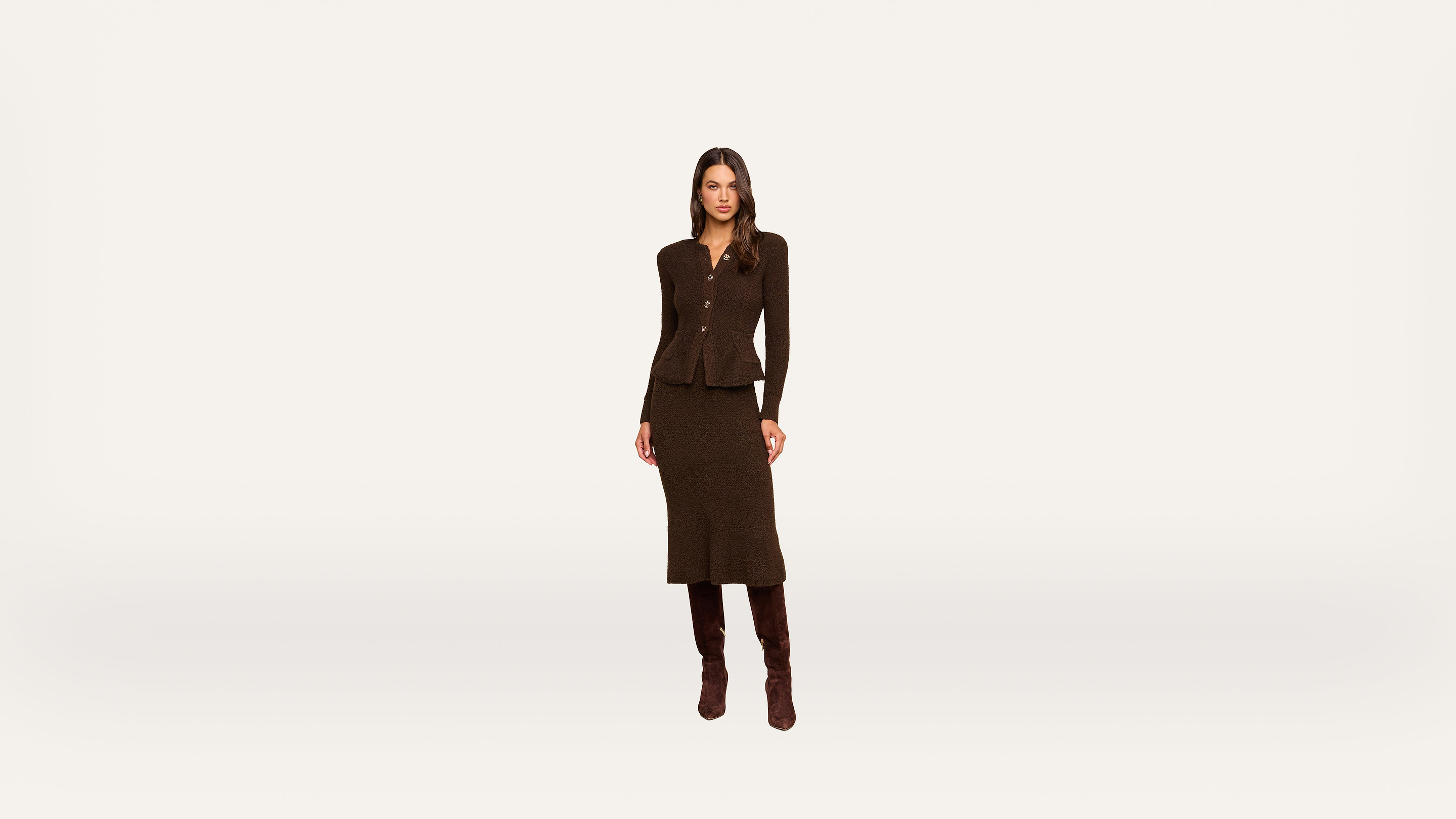 A woman stands against a plain white background, wearing a dark brown long-sleeve top, a matching knee-length skirt, and brown boots.
