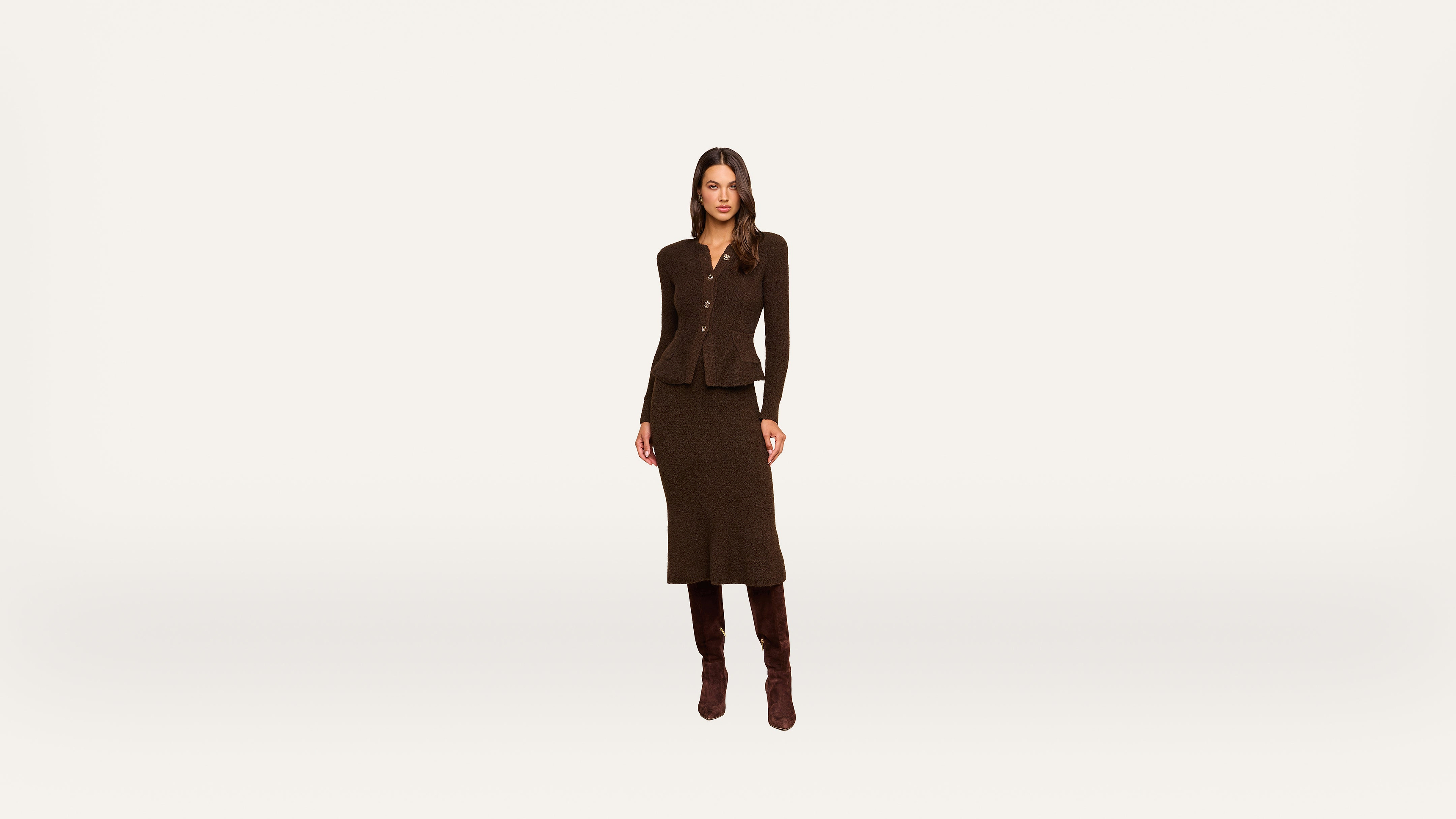 A woman stands against a plain white background, wearing a dark brown long-sleeve top, a matching knee-length skirt, and brown boots.