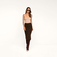 Woman wearing sunglasses, a beige tank top, the Salem Knit Skirt in dark brown, and tall brown boots, standing against a plain beige background.