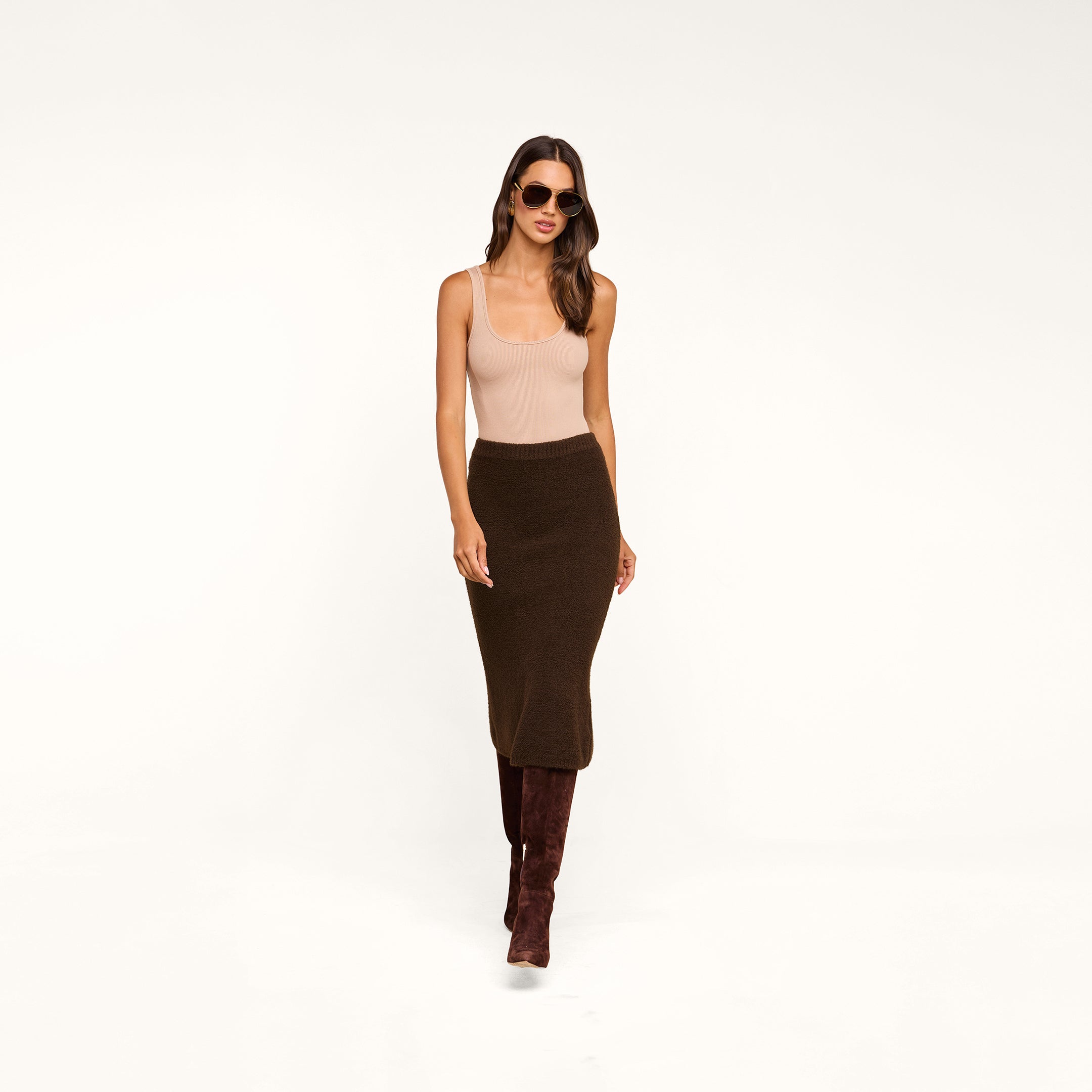 Woman wearing sunglasses, a beige tank top, the Salem Knit Skirt in dark brown, and tall brown boots, standing against a plain beige background.
