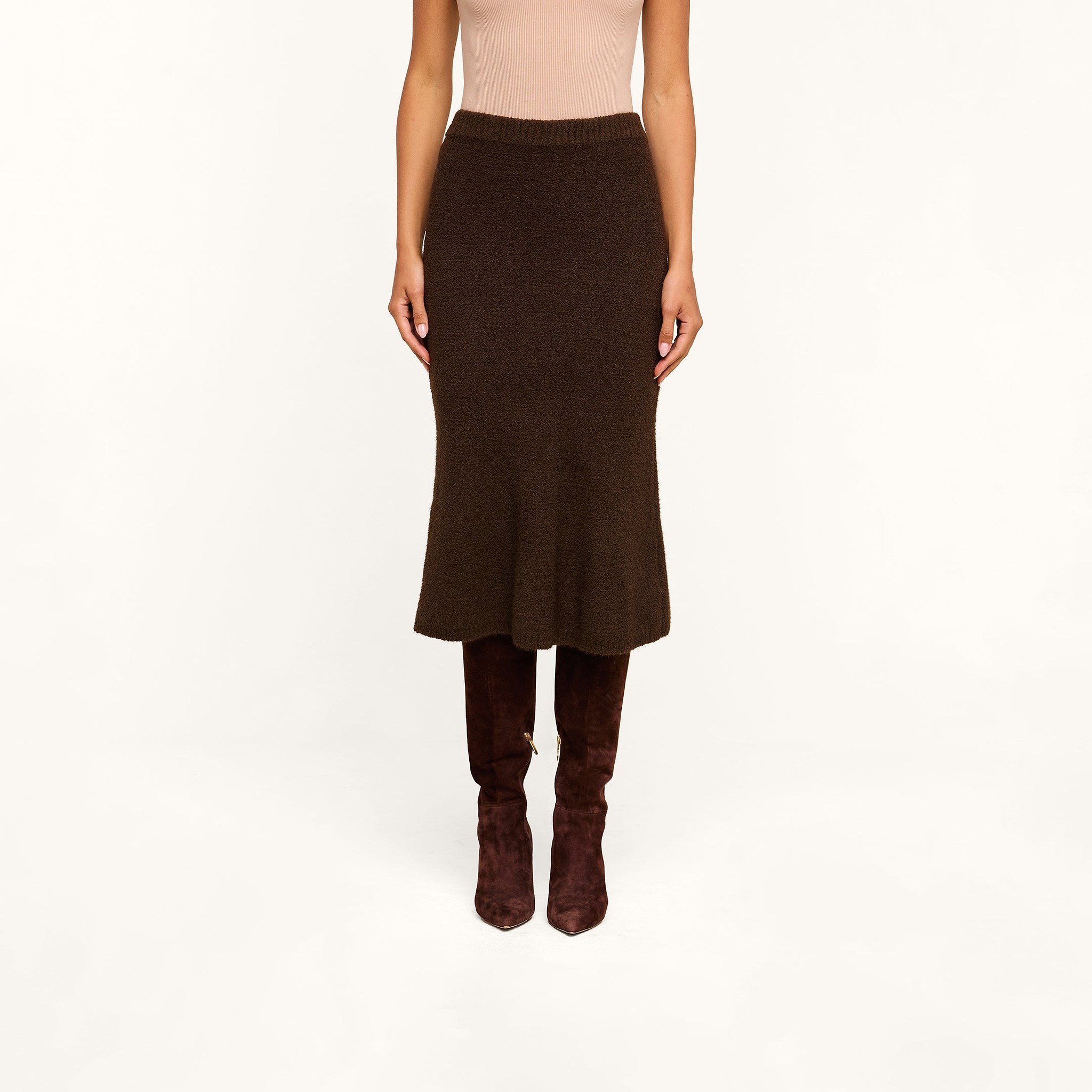 Wearing a light sleeveless top, the person pairs the Salem Knit Skirt in dark brown with brown knee-high suede boots and stands against a plain beige background.