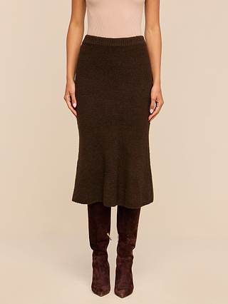 Wearing a light sleeveless top, the person pairs the Salem Knit Skirt in dark brown with brown knee-high suede boots and stands against a plain beige background.