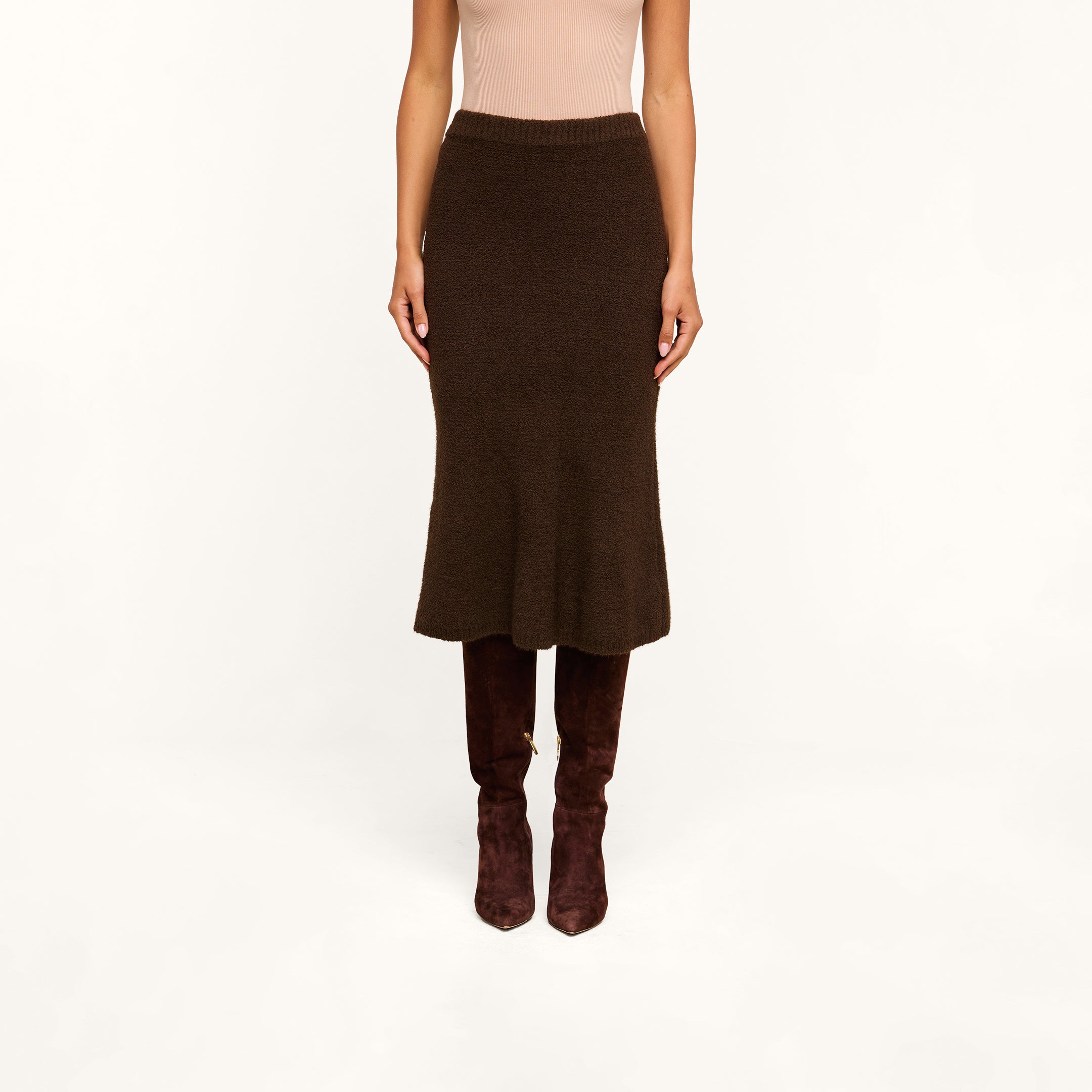 Wearing a light sleeveless top, the person pairs the Salem Knit Skirt in dark brown with brown knee-high suede boots and stands against a plain beige background.