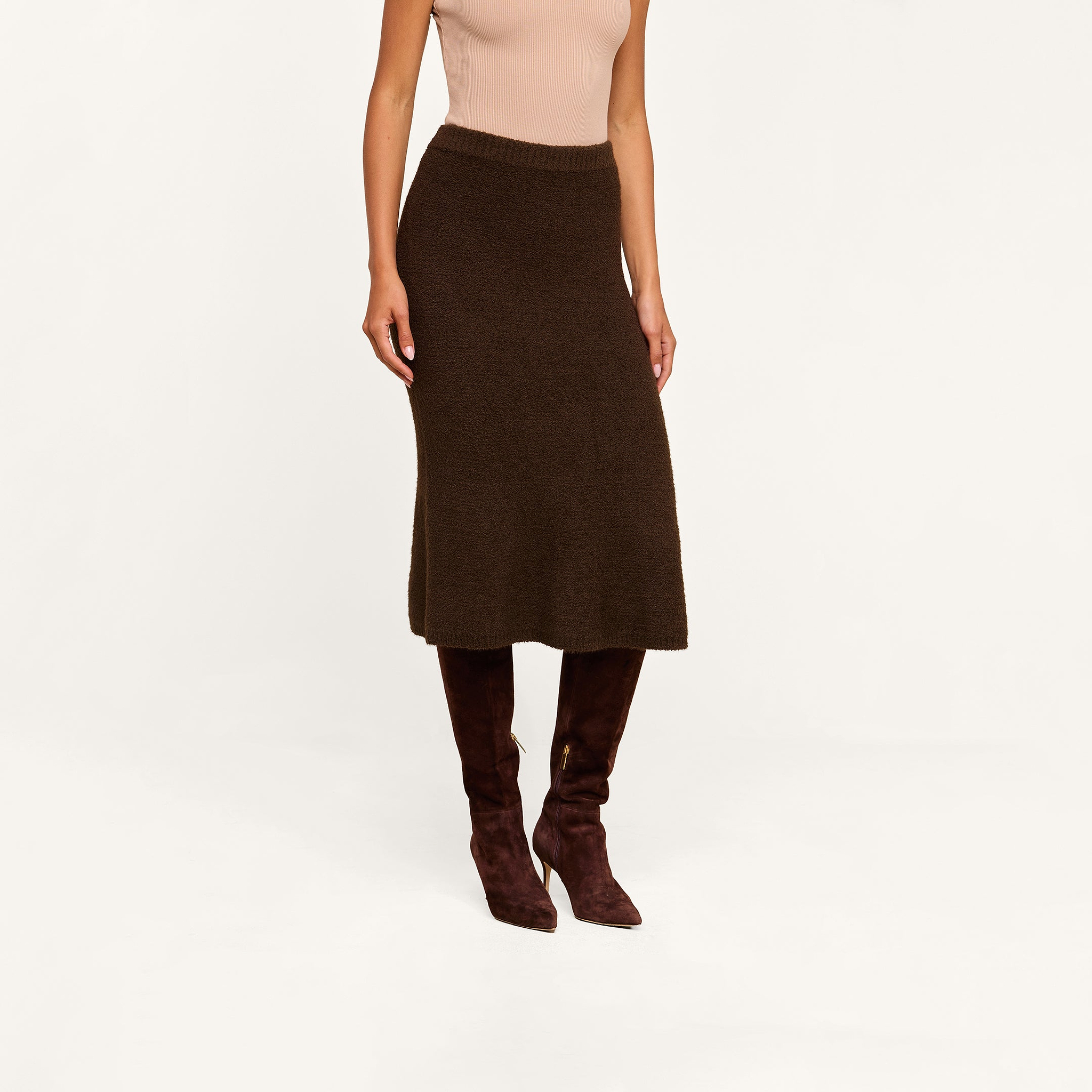 A woman wearing a sleeveless beige top, the Salem Knit Skirt in dark brown, and dark brown knee-high suede boots stands against a beige background.