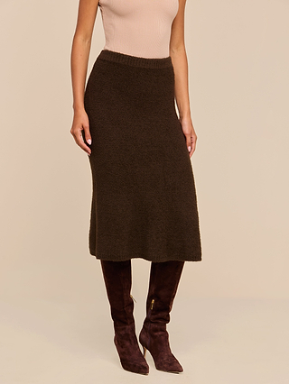 A woman wearing a sleeveless beige top, the Salem Knit Skirt in dark brown, and dark brown knee-high suede boots stands against a beige background.