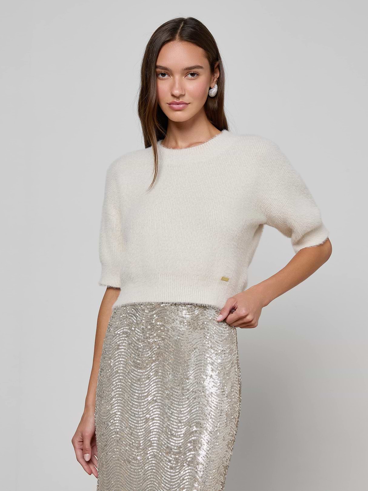 A woman stands against a plain light background, one hand on her hip and a slight smile, wearing the Guinevere Cropped Pullover paired with a high-waisted silver sequin skirt.