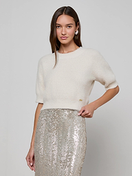 A woman stands against a plain light background, one hand on her hip and a slight smile, wearing the Guinevere Cropped Pullover paired with a high-waisted silver sequin skirt.