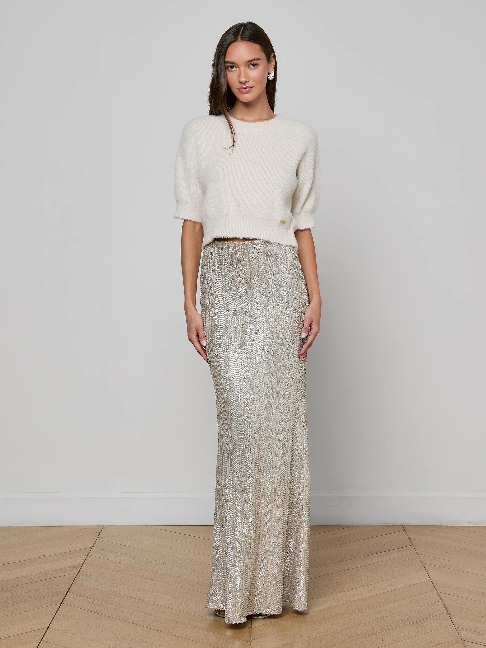 A woman stands wearing the Guinevere Cropped Pullover, a white crewneck sweater in an ultra-soft knit with a cropped silhouette, paired with a long silver sequin skirt. The plain white background highlights her outfit.