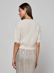 A woman with long brown hair wears the ultra-soft Guinevere Cropped Pullover in white and a silver sequin skirt. She stands facing away, looking over her shoulder against a plain, light background.
