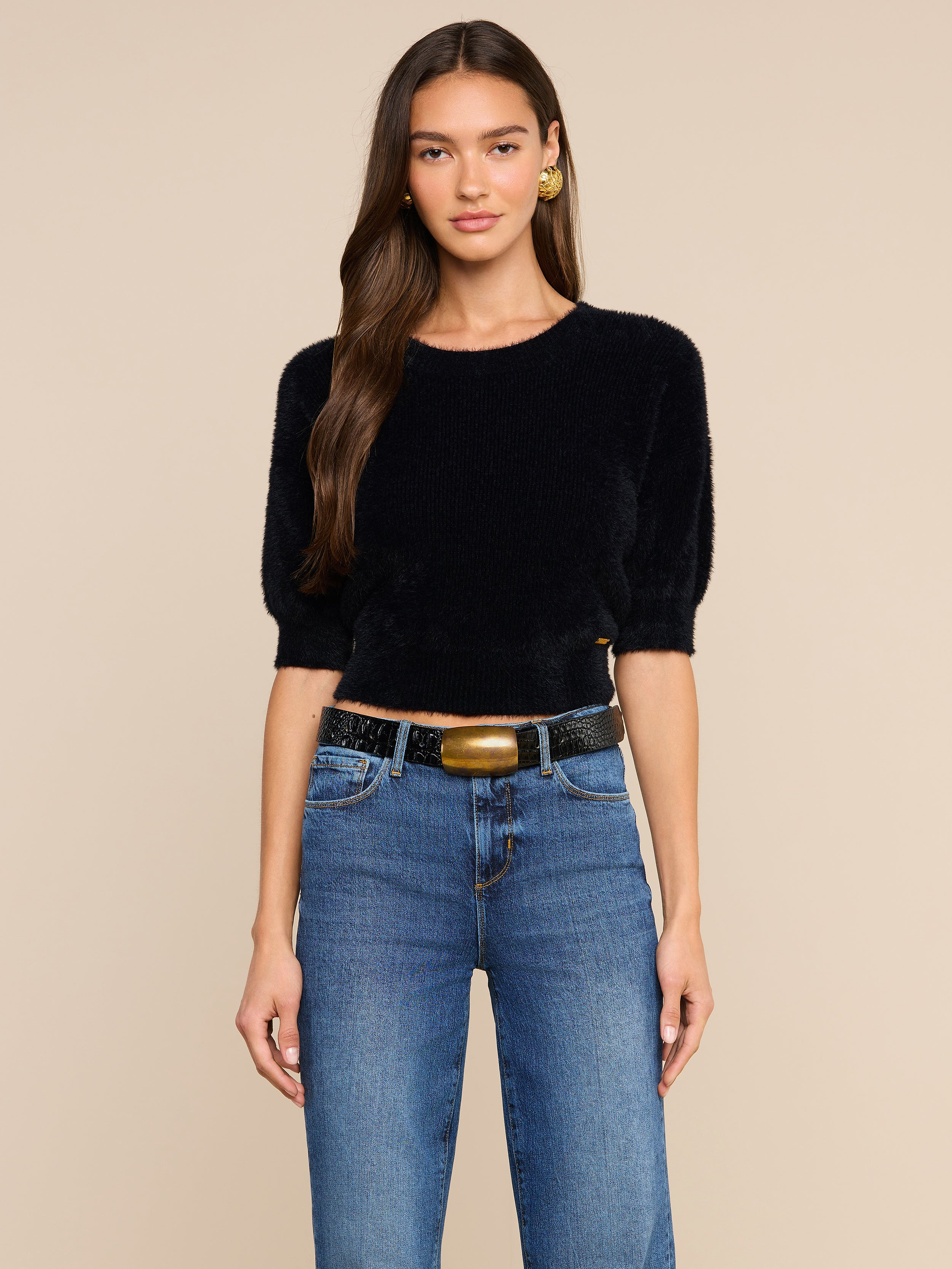 A woman with long brown hair wears the Guinevere Cropped Pullover, a black ultra-soft knit short-sleeve top in a cropped silhouette, styled with blue jeans, a black belt with a gold buckle, and gold earrings, standing against a plain light background.
