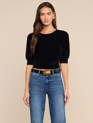 A woman with long brown hair is wearing the Guinevere Cropped Pullover—an ultra soft black knit, short-sleeve fuzzy top with a cropped silhouette—paired with blue jeans, a wide black belt featuring a large gold buckle, and gold hoop earrings, standing against a beige background.