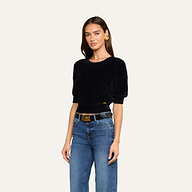 A woman with long brown hair wears the Guinevere Cropped Pullover—a black ultra-soft knit short-sleeve top—paired with blue jeans, a black belt, and gold hoop earrings. She stands against a plain light background, facing slightly to the side.