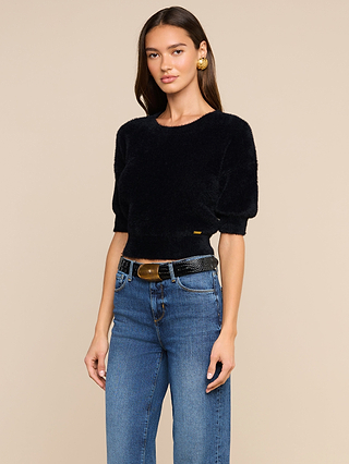A woman with long brown hair wears the Guinevere Cropped Pullover—an ultra-soft, knit fuzzy black short-sleeve crewneck sweater—paired with high-waisted blue jeans and a black belt with a gold buckle, standing against a beige background.