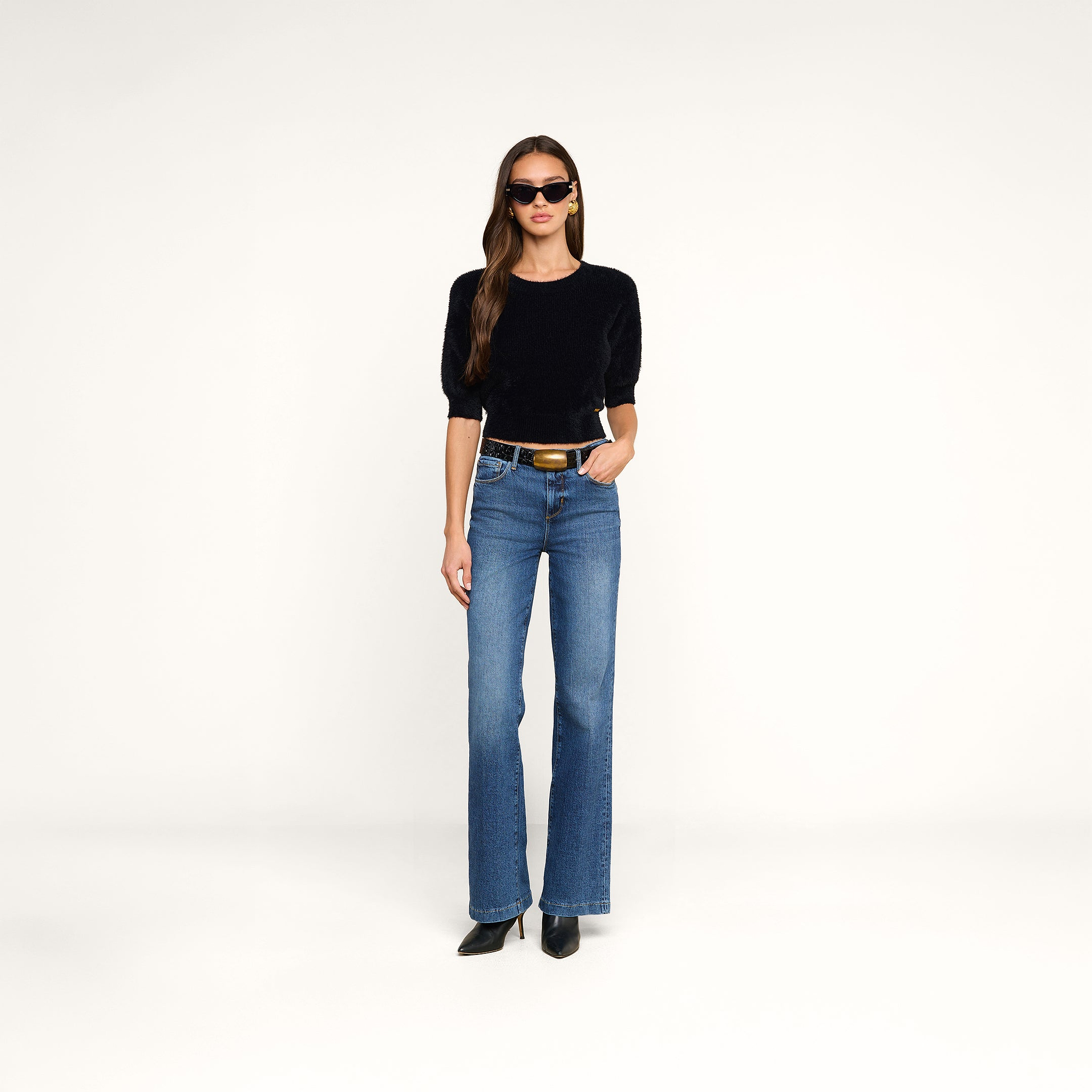 A woman wearing sunglasses, the Guinevere Cropped Pullover—a luxe crewneck sweater with a cropped silhouette—blue high-waisted jeans, a black belt with a gold buckle, and black heeled boots stands against a plain white background.