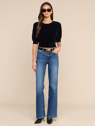 A woman wearing sunglasses, the Guinevere Cropped Pullover—a black ultra soft knit crewneck sweater with a cropped silhouette—blue high-waisted jeans, black heeled boots, and gold earrings, stands against a beige background.