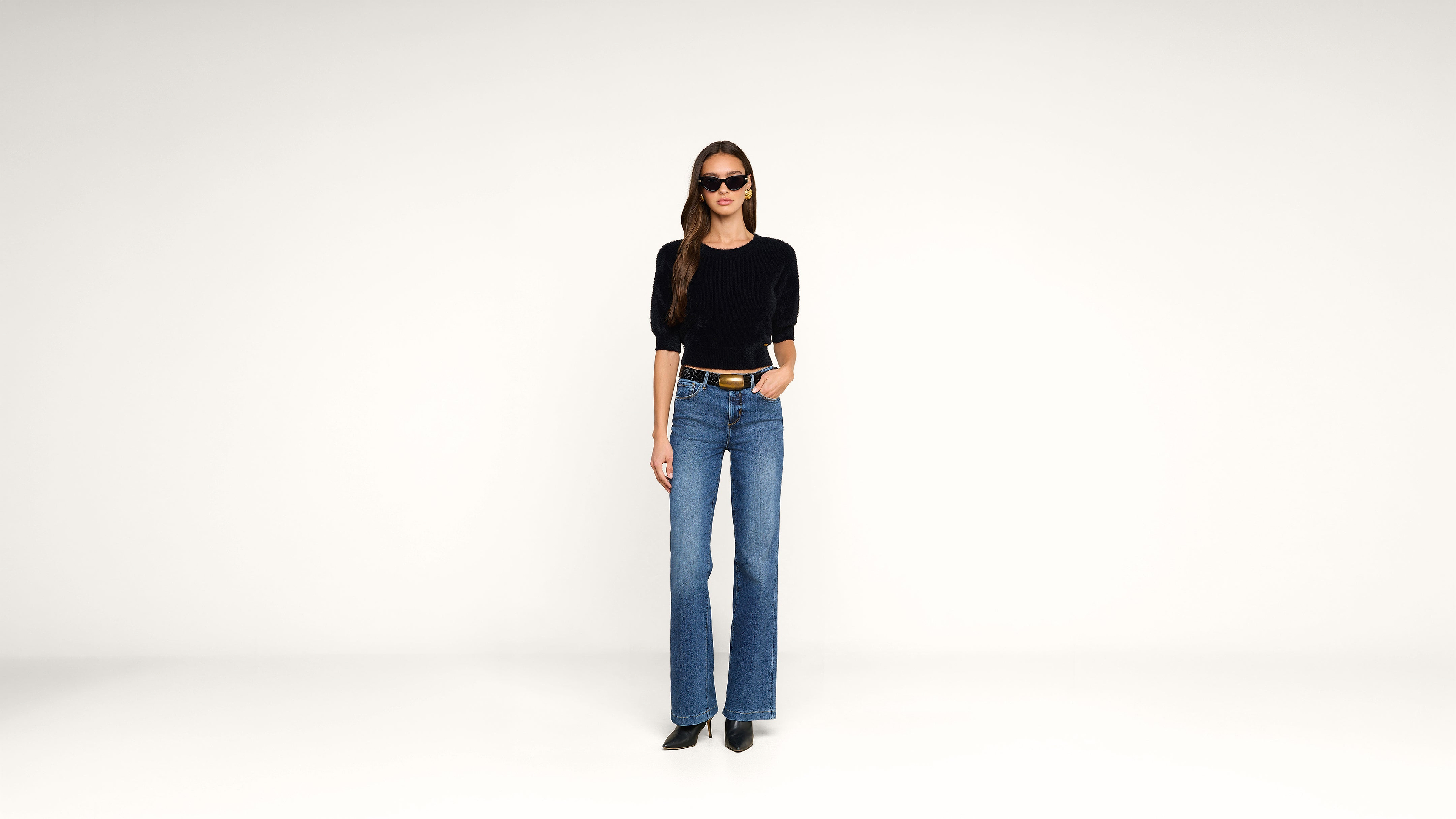 A woman stands against a plain white background, wearing a black top, blue high-waisted jeans, black boots, sunglasses, and a black belt. Her left hand is in her pocket and her hair is down.
