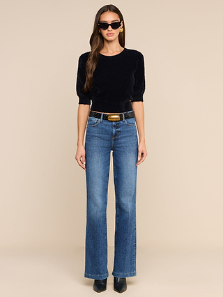 Woman standing against a beige background, wearing the Guinevere Cropped Pullover—an ultra-soft black knit crewneck sweater with a cropped silhouette. She pairs it with blue high-waisted jeans, a black belt, black shoes, and sunglasses. Her long brown hair is loose, and she has a neutral expression.