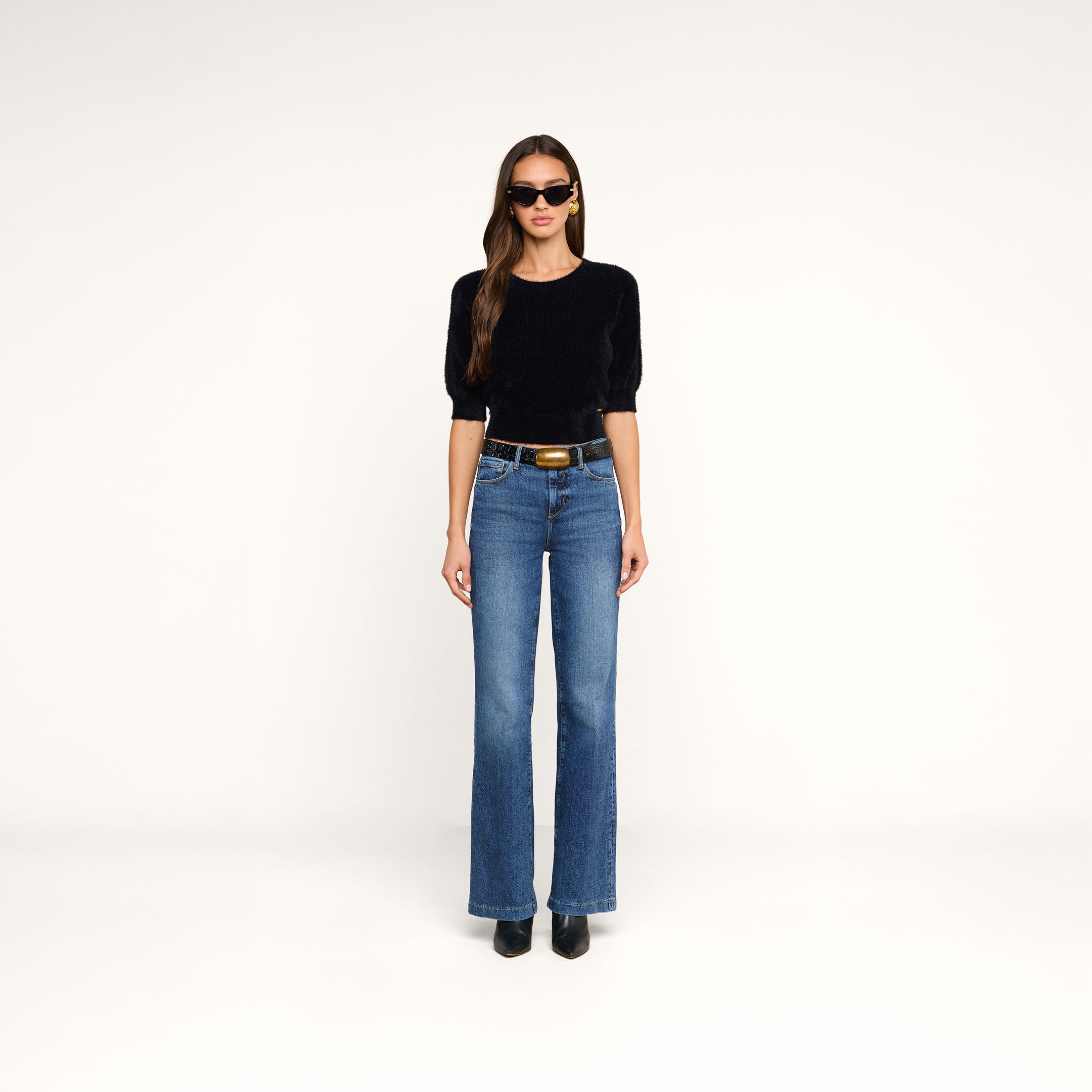 A woman wearing sunglasses, the Guinevere Cropped Pullover—an ultra soft knit black top with a cropped silhouette—blue high-waisted jeans, and black boots stands facing forward against a plain white background.
