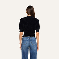 A woman stands facing away, wearing the Guinevere Cropped Pullover—a luxe black crewneck sweater with short sleeves—paired with high-waisted blue jeans. Her long brown hair is down, and her hands rest at her sides against a plain off-white background.