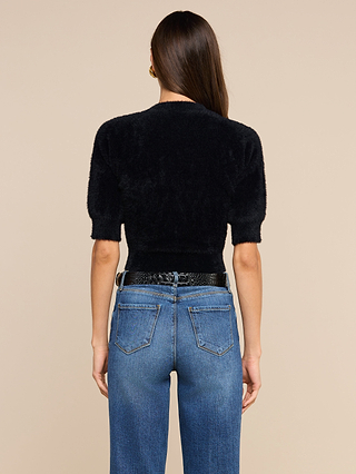 A woman with long brown hair, wearing the Guinevere Cropped Pullover—an ultra soft fuzzy black crewneck sweater—and blue jeans, stands facing away from the camera against a beige background.