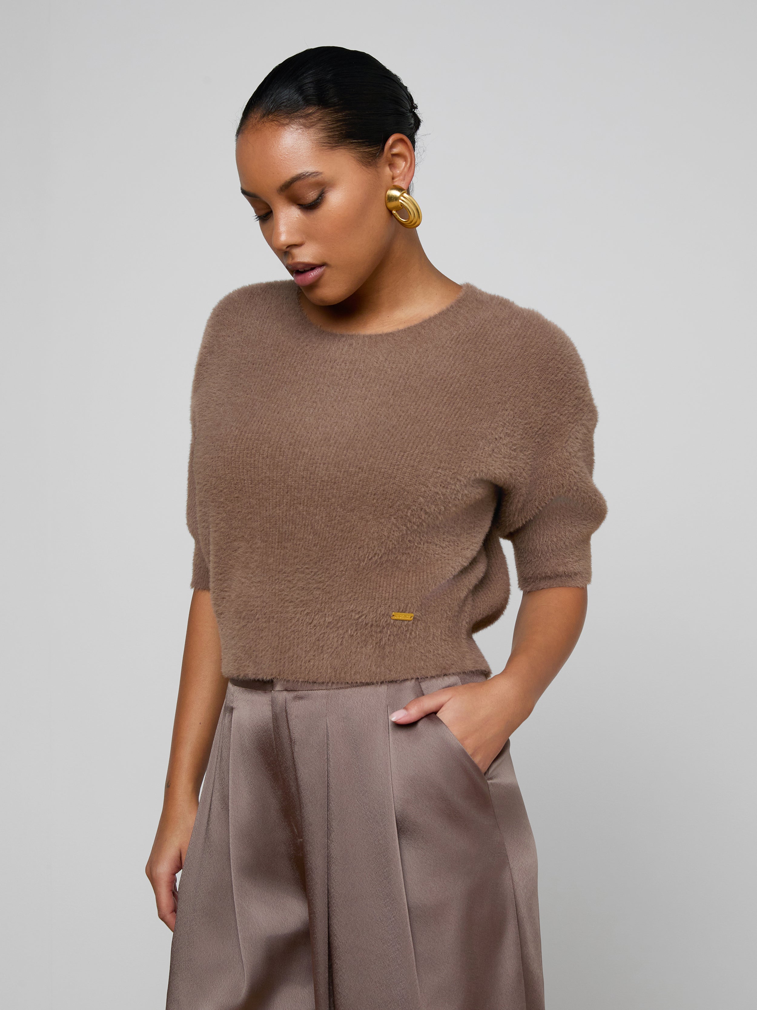 Wearing the Guinevere Cropped Pullover—a brown, ultra-soft crewneck sweater with a cropped silhouette—a woman stands against a plain light background, one hand in her pocket and her gaze cast downward, paired with high-waisted trousers.