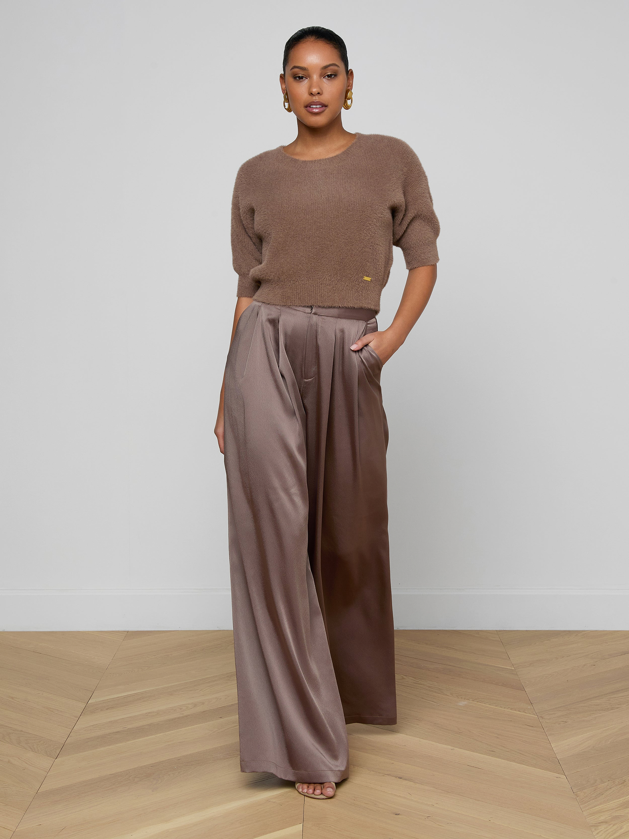 A person stands facing forward against a plain white background, wearing the Guinevere Cropped Pullover—an ultra soft knit brown short-sleeve top—paired with wide-legged, high-waisted brown satin pants with a cropped silhouette, one hand in their pocket.