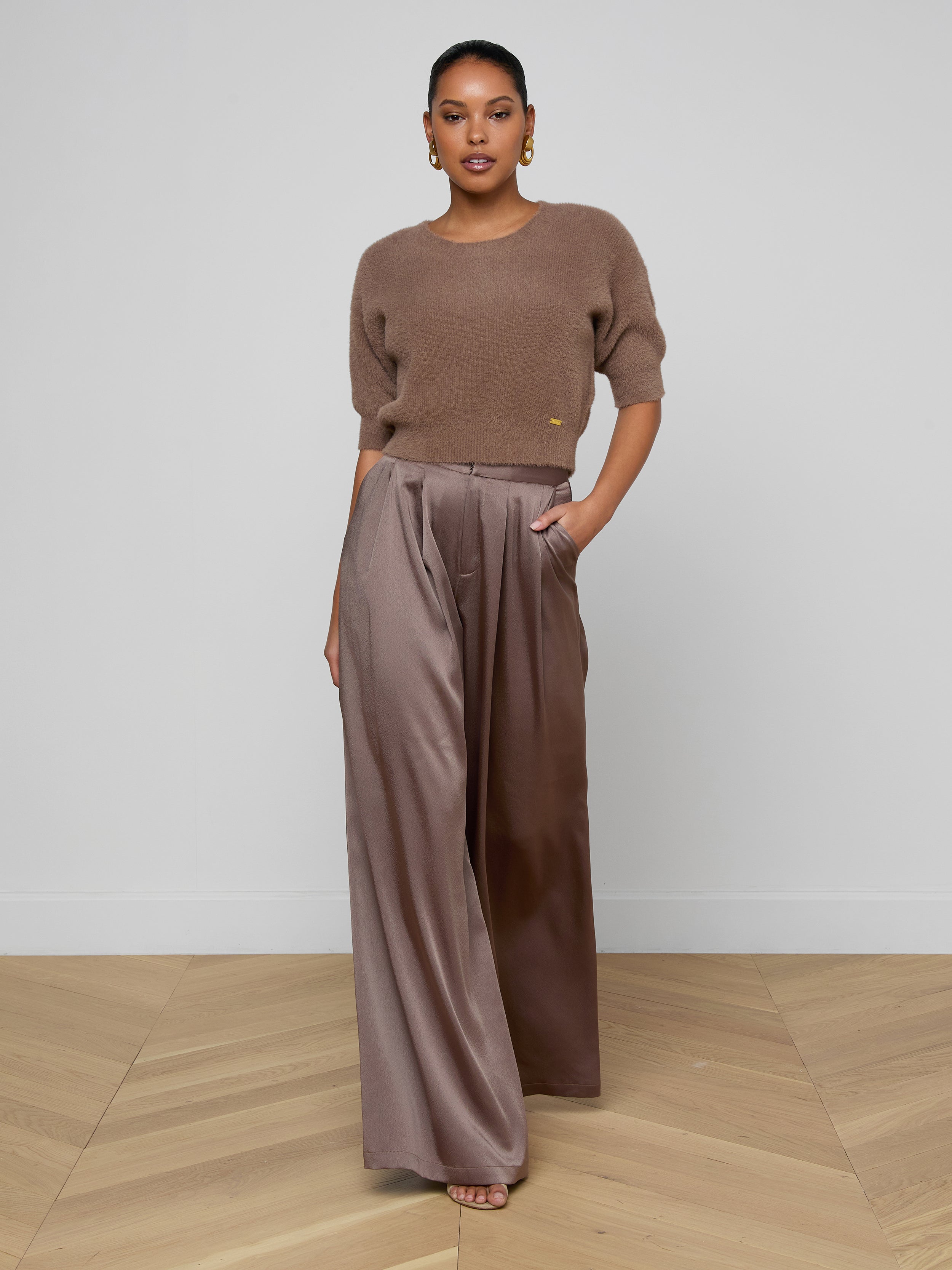 A person stands facing forward against a plain white background, wearing the Guinevere Cropped Pullover—an ultra soft knit brown short-sleeve top—paired with wide-legged, high-waisted brown satin pants with a cropped silhouette, one hand in their pocket.