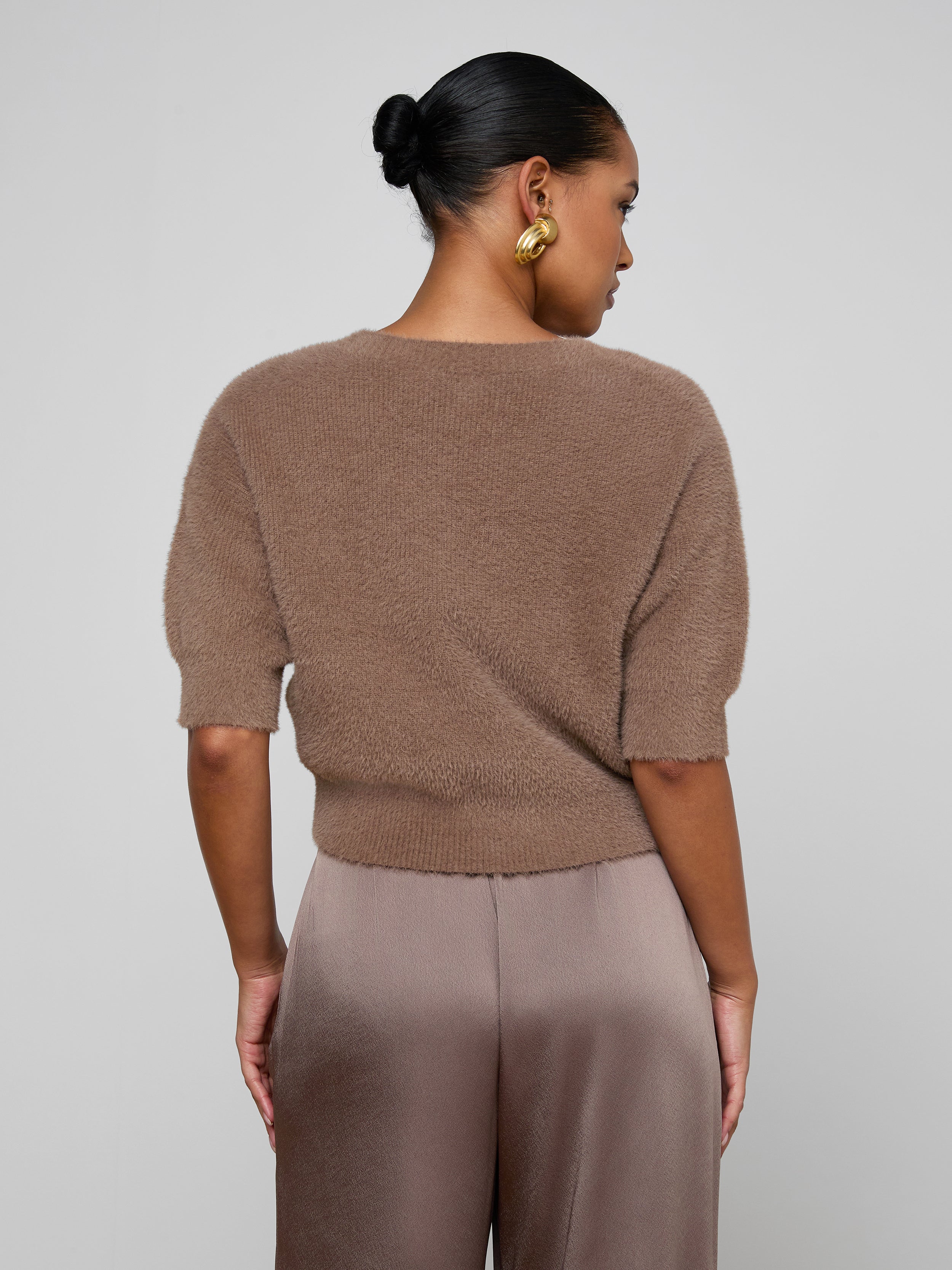 A woman with her hair in a bun and gold hoop earrings is turned away from the camera, revealing the back of the Guinevere Cropped Pullover—a brown ultra-soft knit short-sleeve crewneck sweater with a cropped silhouette—paired with taupe satin pants, set against a plain light background.