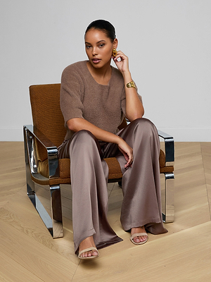 A woman wearing the Guinevere Cropped Pullover sits on a brown chair with chrome arms, paired with loose satin brown pants, nude sandals, a gold bracelet, and large earrings. She looks at the camera, resting one hand on her face.