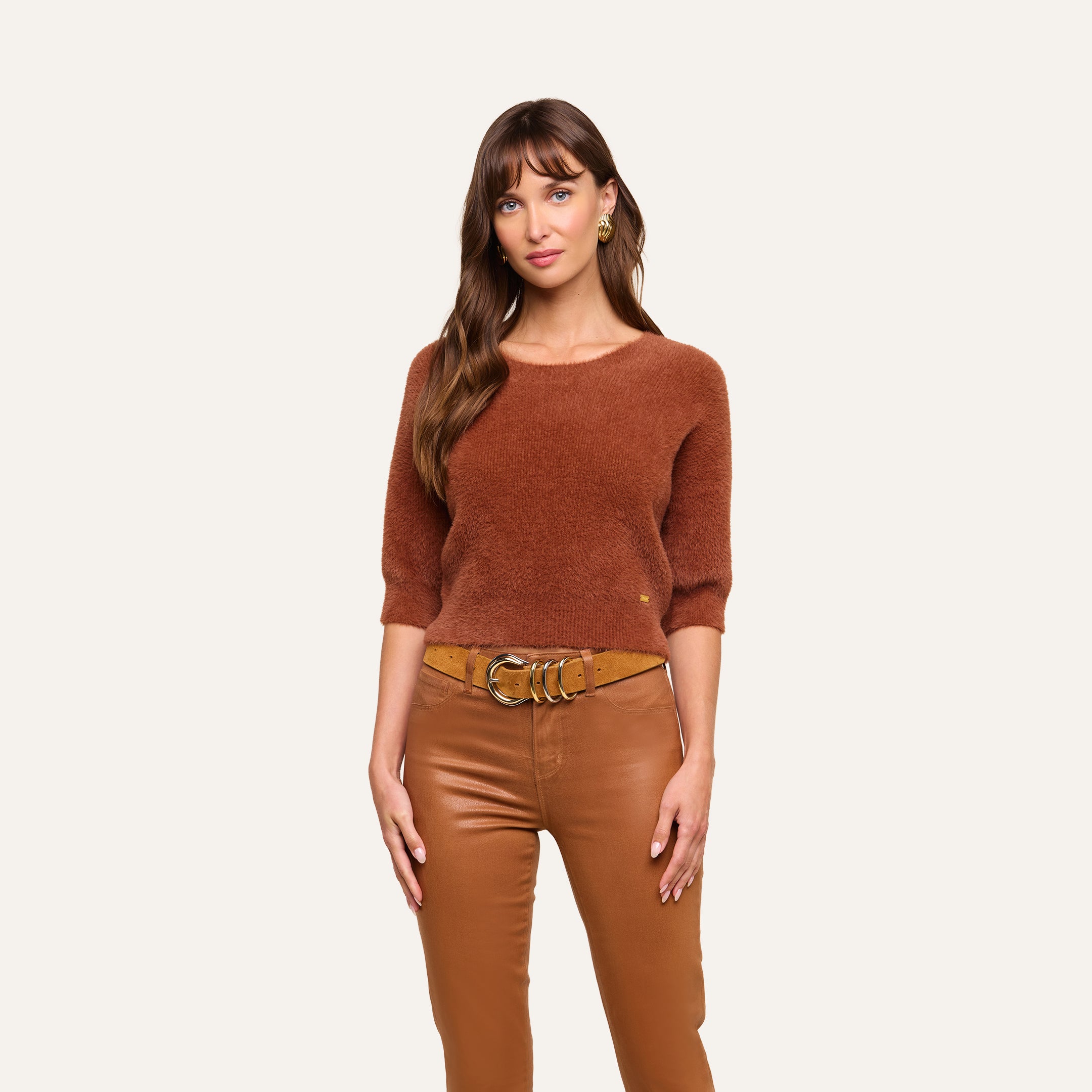 A woman with long brown hair stands against a plain background, wearing the Guinevere Cropped Pullover—an ultra-soft knit, rust-colored crewneck sweater—paired with tan leather pants and a tan belt featuring a large gold buckle. She faces forward with a neutral expression.