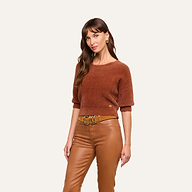 A woman with long brown hair wears the Guinevere Cropped Pullover—a fuzzy brown crewneck sweater with a cropped silhouette—paired with tan leather pants and a tan belt. She accessorizes with gold earrings and stands against a plain light background, looking toward the camera.