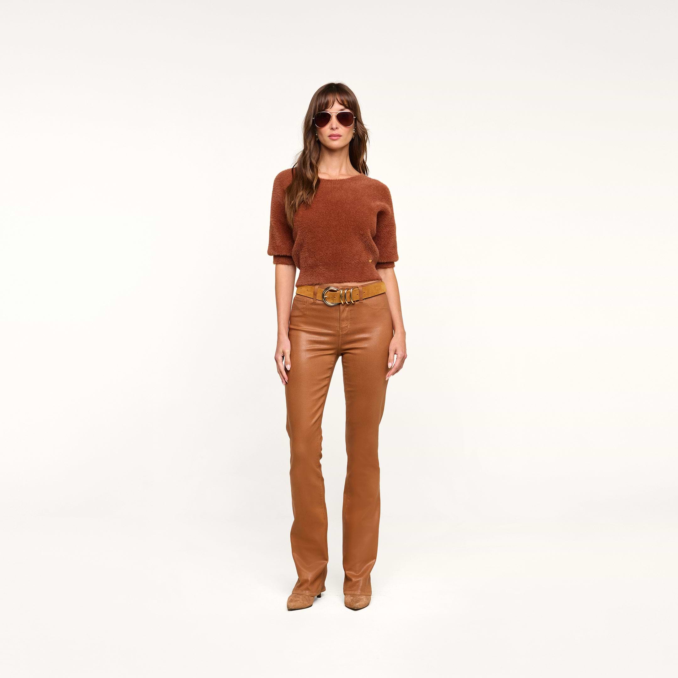 A woman wearing sunglasses and the Guinevere Cropped Pullover—a rust-colored ultra soft knit short-sleeve top with a cropped silhouette—paired with tan leather pants, a tan belt, and tan shoes stands against a plain white background.