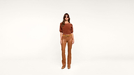 Woman wearing sunglasses, a rust-colored top, tan leather pants, and matching boots stands against a plain white background, facing forward with a neutral expression.