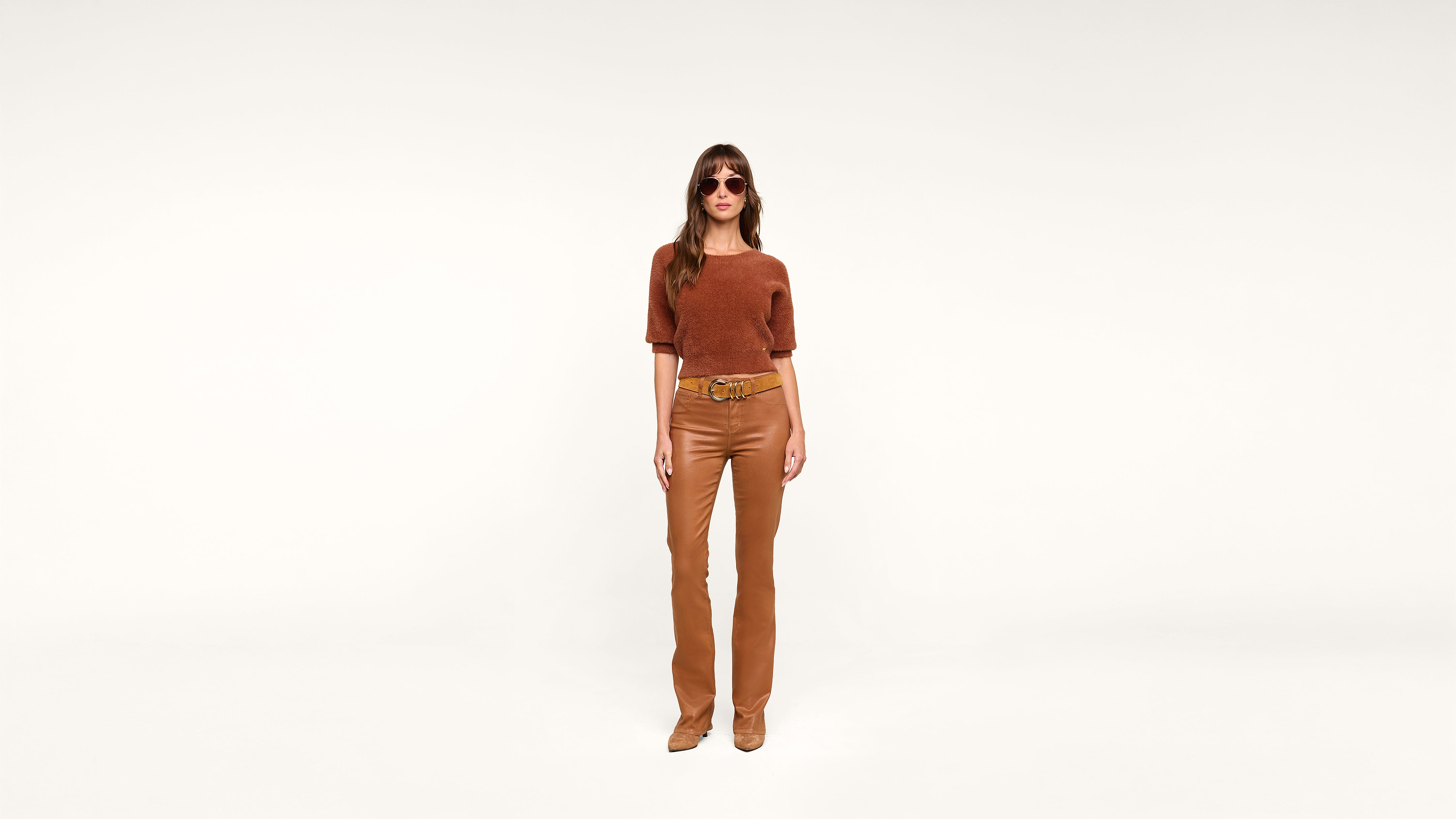 Woman wearing sunglasses, a rust-colored top, tan leather pants, and matching boots stands against a plain white background, facing forward with a neutral expression.
