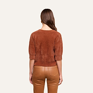A woman with long brown hair stands facing away, wearing the Guinevere Cropped Pullover—an ultra-soft knit crewneck sweater with a cropped silhouette—paired with tan leather pants against a plain light background.