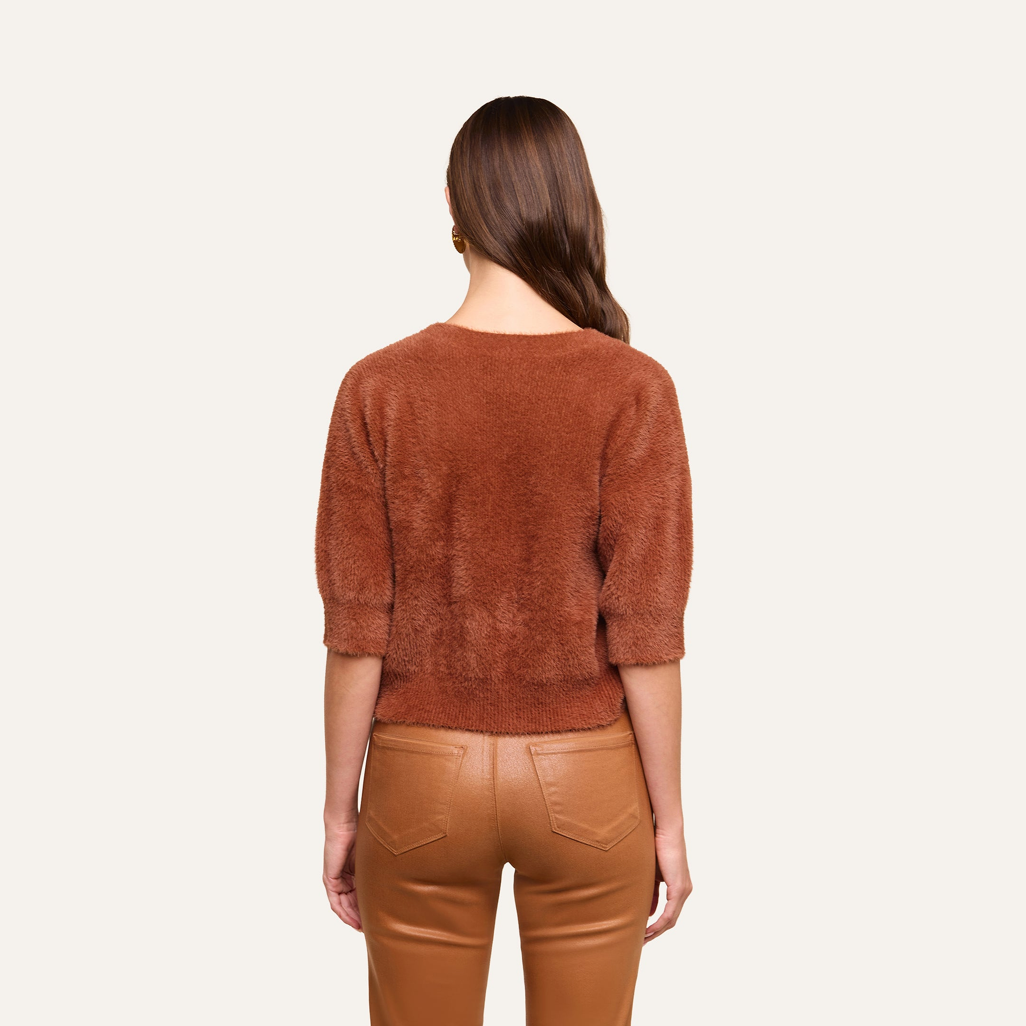 A woman with long brown hair stands facing away, wearing the Guinevere Cropped Pullover—an ultra-soft knit crewneck sweater with a cropped silhouette—paired with tan leather pants against a plain light background.