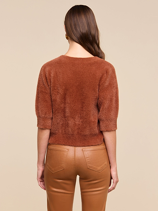 A woman, with loose waves in her brown hair, stands facing away in a Guinevere Cropped Pullover—an ultra-soft, fuzzy brown short-sleeve crewneck sweater—paired with high-waisted tan leather pants. The background is plain beige.