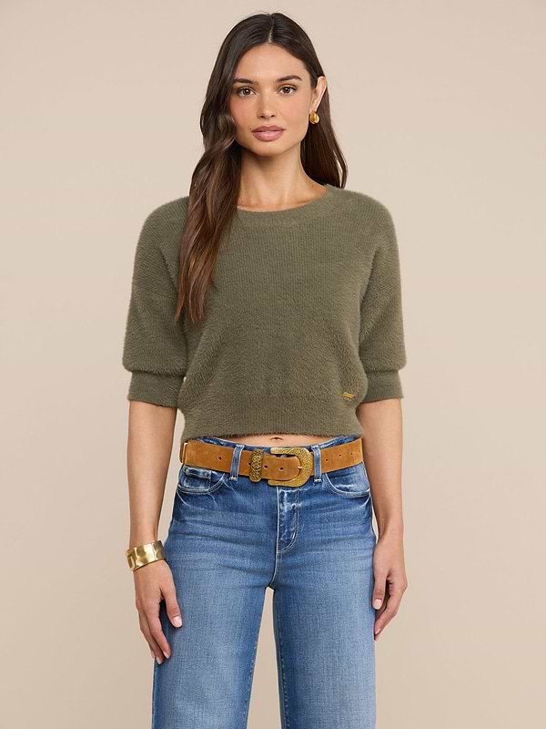 A woman with long brown hair is wearing the Guinevere Cropped Pullover—an olive green crewneck sweater with a cropped silhouette—paired with blue jeans, a tan belt, gold hoop earrings, and a gold bracelet, standing against a plain light background.