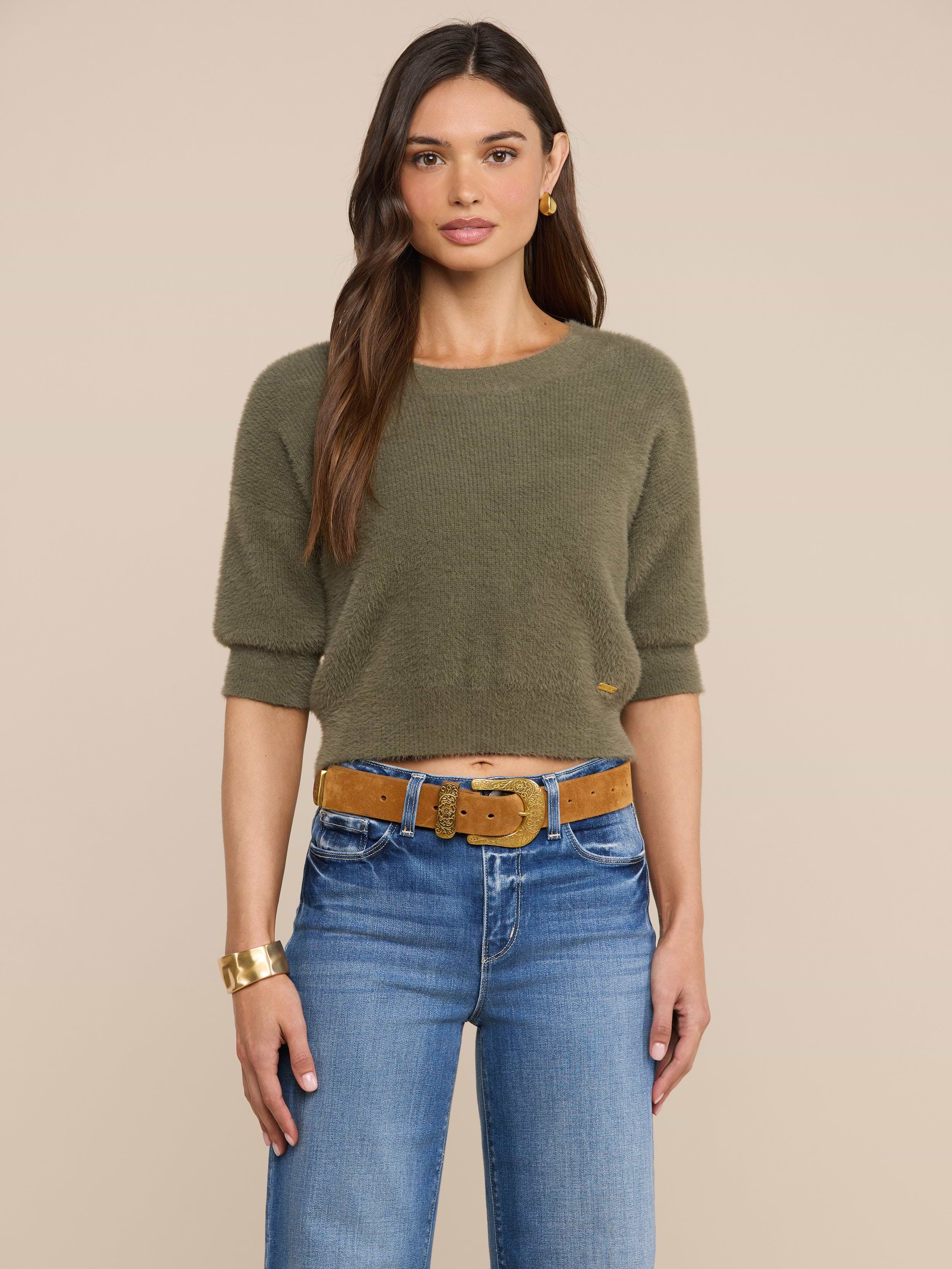A woman with long brown hair is wearing the Guinevere Cropped Pullover—an olive green crewneck sweater with a cropped silhouette—paired with blue jeans, a tan belt, gold hoop earrings, and a gold bracelet, standing against a plain light background.